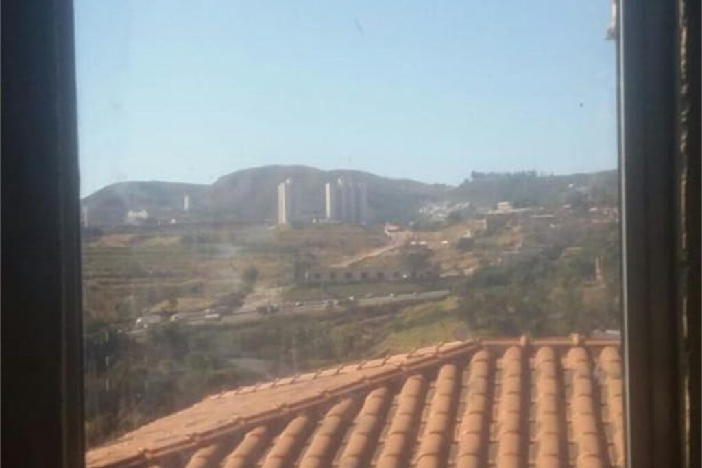 A imagem é a frente de um cartão postal, apresentando uma fotografia nas cores azul, verde e laranja. O dia está claro. Trata-se de uma serra em região da periferia urbana.  A imagem é vista através do vidro de uma janela fechada. Em primeiro plano, na parte inferior, o alto de um telhado colonial. Em segundo plano, um amplo terreno com poucas construções cortado por uma rodovia.  Em terceiro plano, dois prédios altos. Ao fundo, uma serra e o céu azul claro, sem nuvens, ocupando larga faixa superior da imagem. #PraCegoVer #ParaTodosVerem