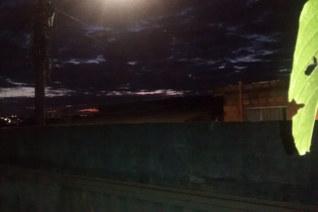 A imagem é a frente de um cartão postal, apresentando uma fotografia em cores, porém pouco iluminada. É início da noite e o céu está nublado. Trata-se da vista do céu sobre um muro. Em primeiro plano, no canto direito, uma única folha verde pende iluminada pelo flash. Em segundo plano, na faixa inferior, está um muro alto no qual se vê apenas a parte superior de um portão de garagem. Em terceiro plano, através do muro, há um poste aceso e o topo de algumas casas. Ao fundo, o céu está escuro, mas é possível avistar muitas nuvens e um resquício de claridade no horizonte. #PraCegoVer #ParaTodosVerem