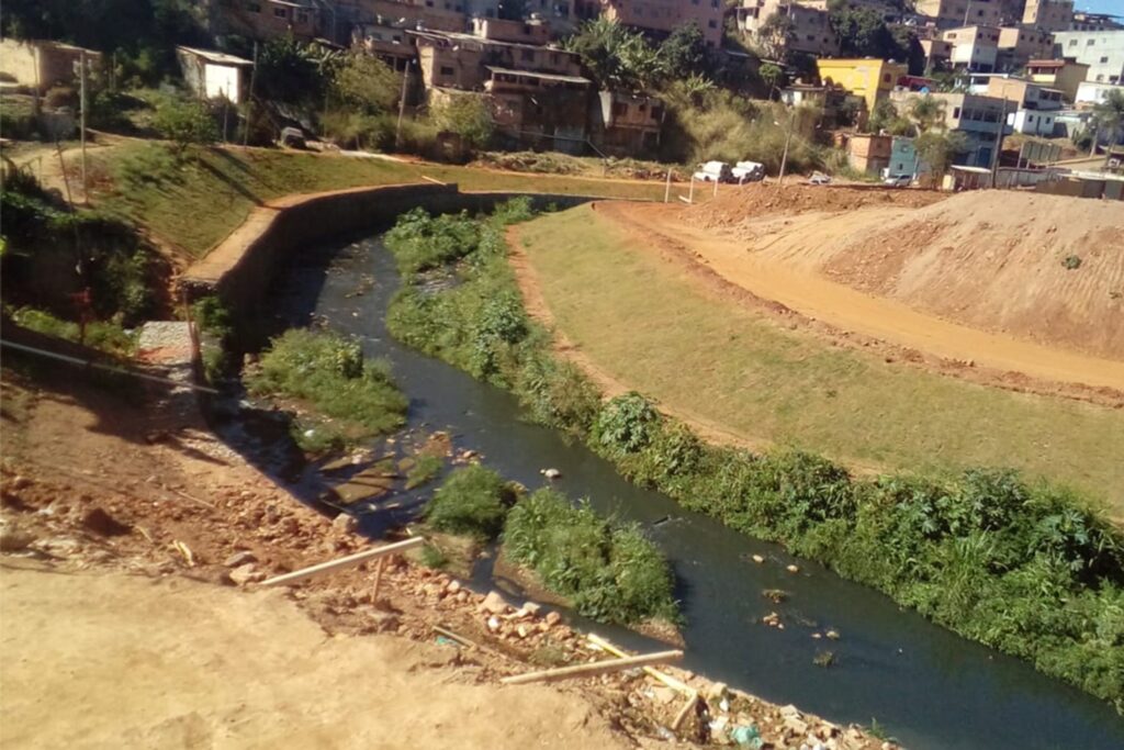 A imagem é a frente de um cartão postal, apresentando uma fotografia onde predominam tons de verde e terrosos. O dia está claro. Trata-se da curva de um córrego que passa em meio a área urbana. Em primeiro plano, bem no centro da imagem, em destaque, o córrego tem águas esverdeadas e turvas e parece haver lixo em suas águas. Na margem esquerda, está contido por muro de concreto. Em sua margem direita, há uma vegetação baixa e, em segundo plano, um terreno com um grande monte de terra e o início de uma via de terra. Ao fundo da imagem, no faixa superior, um aglomerado de casas populares. #PraCegoVer #ParaTodosVerem