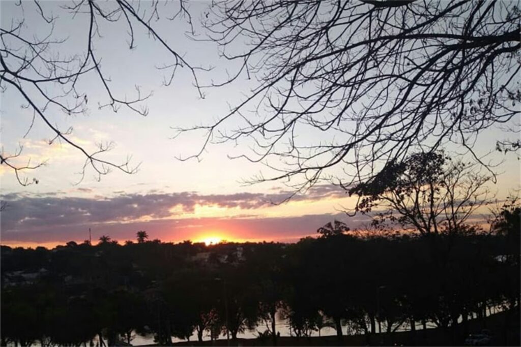 A imagem é a frente de um cartão postal, apresentando uma fotografia em cores, com destaque para tons violeta e o contraste entre luz e sombra. É fim de tarde. Trata-se de visão panorâmica do pôr-do-sol visto de uma lagoa.  Em primeiro plano, na faixa superior, galhos secos à sombra emolduram o quadro. Em segundo plano, na faixa inferior, uma fileira de árvores do mesmo tipo, também à sombra, deixa entrever parte de uma lagoa. Ao fundo, no centro da imagem, em destaque, o sol se põe quase por completo, tingindo as nuvens no horizonte em tons de vermelho e lilás. #PraCegoVer #ParaTodosVerem 