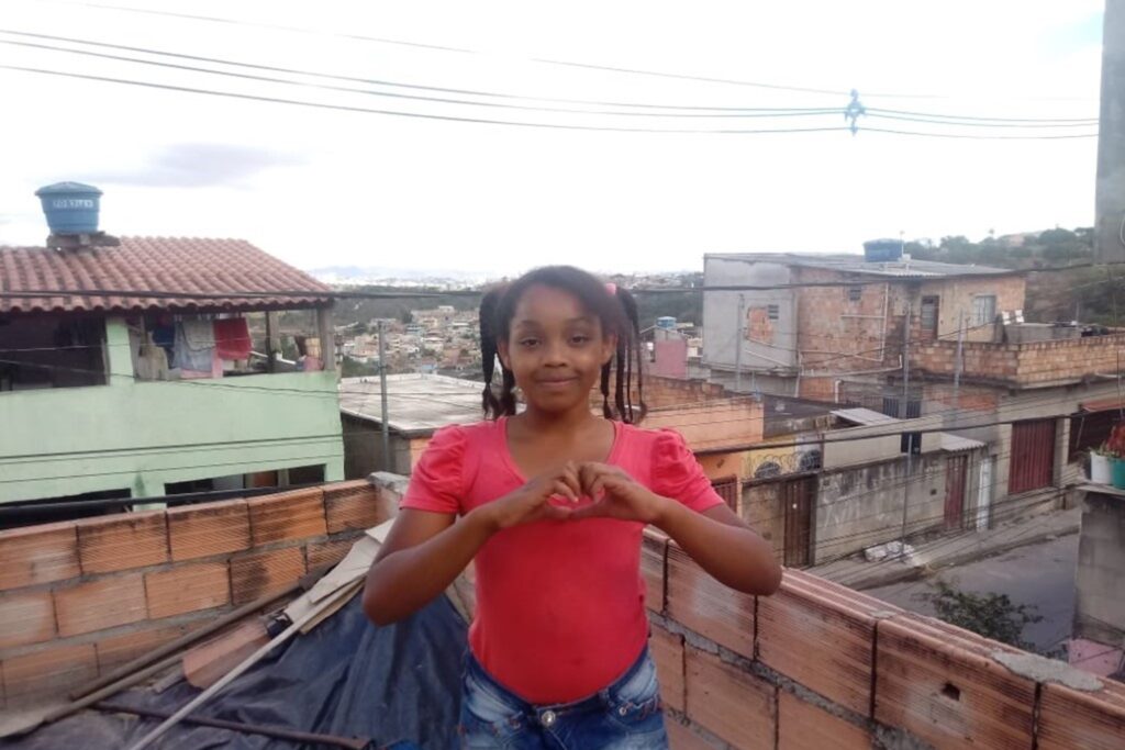 A imagem é a frente de um cartão postal, apresentando uma fotografia colorida em que predominam tons alaranjados. O dia está nublado. Trata-se de uma criança sorrindo. Em primeiro plano, uma menina negra, com cabelos presos e trançados, blusa vermelha e shorts jeans, sorri para a câmera e faz com as mãos um coração. Ela está no alto de uma laje com muros baixos em tijolo aparente. Em segundo plano, trecho da rua e as casas vizinhas. Bem ao fundo, se vê outras casas. Na faixa superior, céu claro, coberto de nuvens e alguns fios de energia. #PraCegoVer #ParaTodosVerem