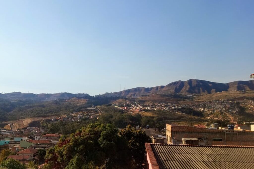 A imagem é a frente de um cartão postal, apresentando uma fotografia onde predominam tons de verde e azul. O dia está claro. Trata-se de uma vista panorâmica de periferia urbana. Em primeiro plano, se vê o topo de uma árvore no limite inferior do enquadramento. Ao lado direito da árvore, parte do telhado de uma casa. Um pequeno aglomerado de casas forma uma fina mancha urbana que se estende do primeiro plano da imagem até a Serra do Curral, com suas curvas em destaque ao fundo, marcando toda a linha do horizonte e o limite do céu limpo, azulado e sem nuvens. #PraCegoVer #ParaTodosVerem