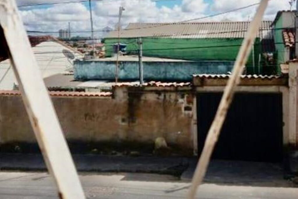 A imagem é a frente de um cartão postal, apresentando uma fotografia em cores, com destaque para tons claros, de azul e verde. O dia está claro. Trata-se da casa do vizinho de frente vista pela janela. Em primeiro plano, fora do foco, se vê duas barras de esquadrias metálicas brancas e finas da janela. Em segundo plano, através delas, observa-se um pequeno trecho da rua de asfalto e um muro com portão preto de garagem. Em terceiro plano, por trás do muro, estão terraços de casas nas cores azul, verde e branca. Fios de energia cruzam a imagem na horizontal. Bem ao fundo, uma pequena faixa de céu repleto de nuvens. #PraCegoVer #ParaTodosVerem