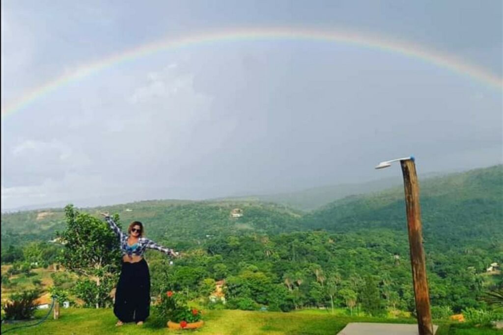 A imagem é a frente de um cartão postal, apresentando uma fotografia em que predominam os tons de verde e cinza. O dia está nublado. Trata-se de um grande arco-íris sobre uma mata. Em primeiro plano, na faixa inferior, à esquerda, há uma mulher de óculos escuros, camisa xadrez azul e saia longa preta posando para a câmera de braços abertos. À direita, há uma ducha presa a um mastro de madeira. Ao fundo, mares de morros, com densa vegetação em tons de verde. Na larga faixa superior da imagem, um estreito e comprido arco-íris atravessa o quadro da extremidade esquerda à direita sobre um céu cinza.  #PraCegoVer #ParaTodosVerem  