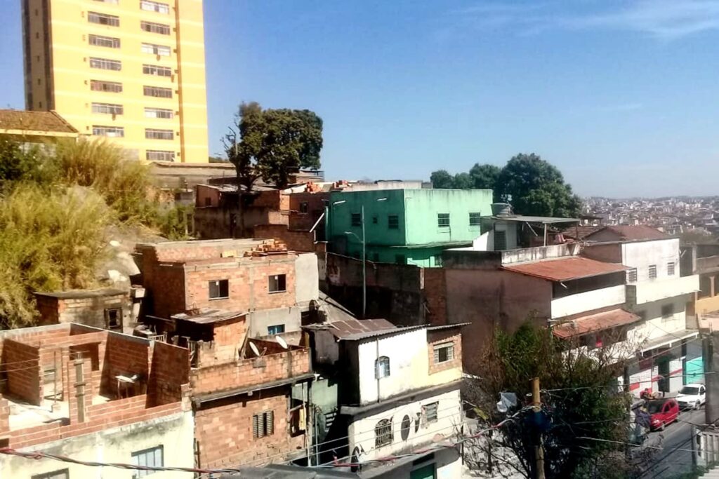 A imagem é a frente de um cartão postal, apresentando uma fotografia em cores, com predomínio do azul e de tons terrosos. O dia está claro. Trata-se de uma rua residencial vista de cima. Em primeiro plano, uma esquina com ladeira íngreme corta dois quarteirões com residências de dois e três andares. O quarteirão da direita tem casas em construção. No canto inferior direito, dois carros, um branco e um vermelho, na rua estacionados. Em segundo plano, no canto superior esquerdo, destaca-se um prédio amarelo, de muitos andares, do qual a imagem corta o topo. Bem ao fundo, do lado direito, se vê a continuação da cidade e em toda parte superior, larga faixa de céu azul sem nuvens. #PraCegoVer #ParaTodosVerem