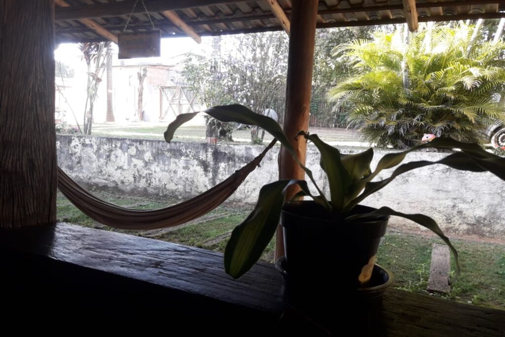 A imagem é a frente de um cartão postal, apresentando uma fotografia onde predomina o verde e tons de madeira. É dia. Trata-se do jardim de uma casa. Em primeiro plano, parapeito de uma janela de madeira, à sombra, com vaso de plantas. Em segundo plano há um pilar, também de madeira, no qual se prende uma rede de descanso que se estende até o lado esquerdo do quadro. Em terceiro plano, na faixa superior da imagem, a extremidade interna de um telhado colonial. Em quarto plano, um gramado e um muro baixo. Em quinto plano, um terreno em nível mais elevado, com portão bege e pequenas árvores e arbusto, atrás do qual é possível ver parte da roda de um carro. #PraCegoVer #ParaTodosVerem