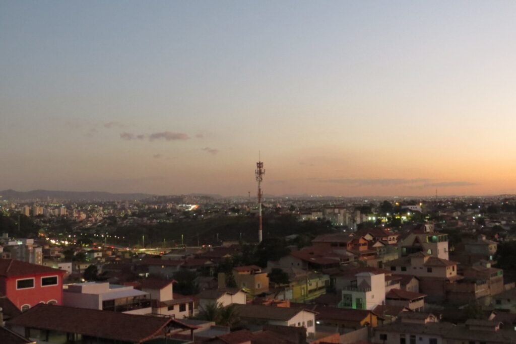 A imagem é a frente de um cartão postal, apresentando uma fotografia em que predominam tons claros de azul acinzentado e laranja e tons escuros de marrom e verde. Está anoitecendo. Trata-se de uma visão panorâmica de paisagem urbana. Em primeiro plano, na faixa inferior, várias casas de até dois andares com telhados coloniais. Alguns postes e luzes das casas já estão acesos. Em segundo plano, no centro da imagem, destaca-se uma torre alta e fina nas cores branca e laranja. Ao fundo, a paisagem urbana se estende até o fim do horizonte. No canto esquerdo, ao fundo, se vê parte de uma serra. Ocupando uma larga faixa superior, o céu já sem a luz direta do sol, mas ainda claro, tem tons de azul, cinza e laranja. #PraCegoVer #ParaTodosVerem