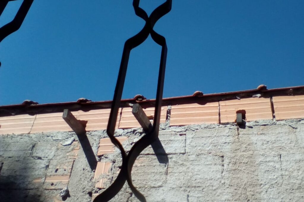 A imagem é a frente de um cartão postal, apresentando uma fotografia em tons de azul, terra e cinza. É dia. Trata-se do céu visto de dentro de casa.  A imagem está angulada de baixo para cima e foi feita através de uma janela. Em primeiro plano, uma grade decorativa. Em segundo plano, do lado de fora, um muro de tijolos aparentes, parcialmente rebocado com cimento, cobre toda a metade inferior da imagem.  Na metade superior, céu sem nuvens em cor azul intenso. #PraCegoVer #ParaTodosVerem