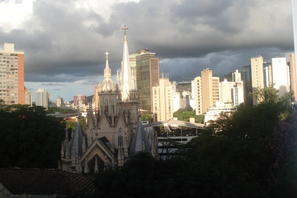 A imagem é a frente de um cartão postal, apresentando uma fotografia colorida em que predomina o contraste entre luz e sombra. O dia está nublado. Trata-se de torres de uma igreja em paisagem urbana. Em primeiro plano, na faixa inferior, a copa de algumas árvores e um telhado encobertos de sombra. Em segundo plano, no centro da imagem, em destaque, a parte superior de uma igreja, com várias torres pontiagudas com cruzes no topo. A igreja está parcialmente encoberta pela sombra e somente duas torres mais altas estão iluminadas pelo sol. Ao fundo, em toda a faixa central, prédios altos. Na faixa superior, muitas nuvens carregadas. #PraCegoVer #ParaTodosVerem