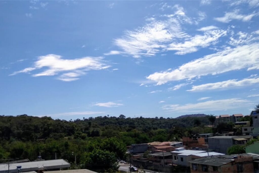 A imagem é a frente de um cartão postal, apresentando uma fotografia em cores, predominando o azul celeste. É dia. Trata-se de visão panorâmica sobre uma mata. Em primeiro plano, à direita, algumas casas são vistas de cima. Em segundo plano, na faixa inferior à esquerda, uma grande mata em tom verde escuro se estende até o fundo do canto direito. Ao fundo, o céu azul, muito iluminado e com poucas nuvens, ocupa a maior parte do enquadramento. #PraCegoVer #ParaTodosVerem