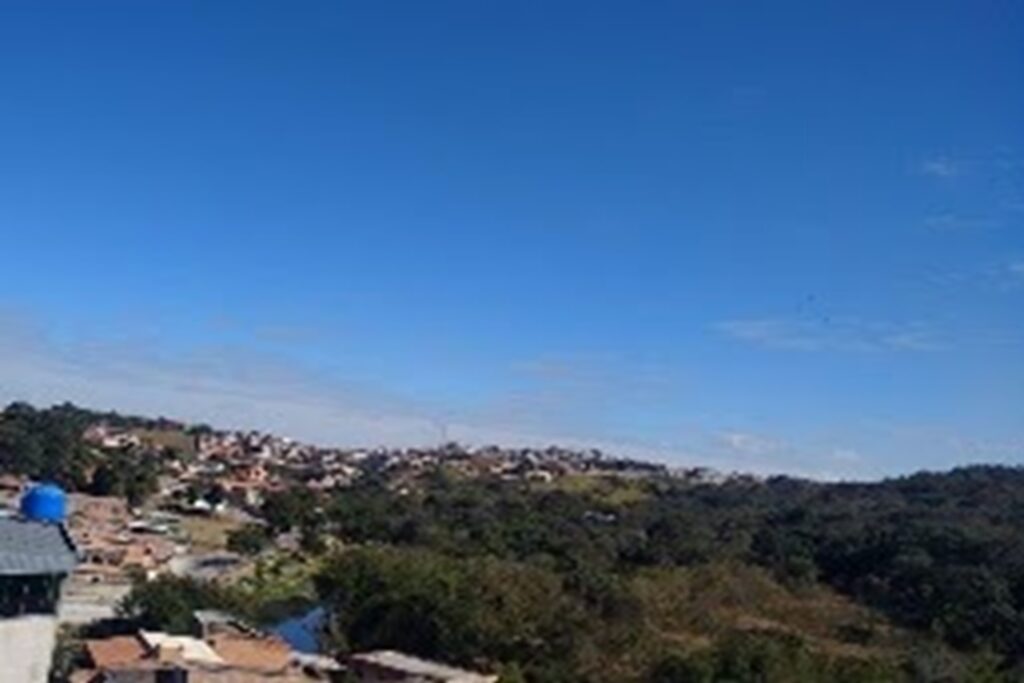 A imagem é a frente de um cartão postal, apresentando uma fotografia colorida em que predominam o azul e o verde. É dia. A foto levemente angulada para cima registra o céu sobre paisagem verde. Em primeiro plano, na faixa inferior, vê-se a copa das árvores em mata fechada e uma faixa ocupada por casas que se inicia em primeiro plano, no canto esquerdo, e se estende até o fundo da imagem, no horizonte. Ocupando a maior parte do quadro, céu muito azul e limpo. #PraCegoVer #ParaTodosVerem