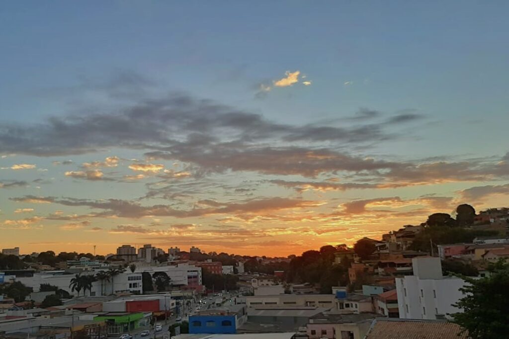 A imagem é a frente de um cartão postal, apresentando uma fotografia em que predominam tons de azul e amarelo. O dia está no crepúsculo. Trata-se de uma paisagem urbana em visão panorâmica, com destaque para o céu. Em primeiro plano, na parte superior da imagem, o céu é azul com poucas nuvens e, na faixa inferior direita, há vários prédios. Em segundo plano, na faixa inferior, a mancha urbana é cortada por uma avenida larga com muitos carros e outros prédios ocupam o canto inferior esquerdo da imagem. Ao fundo, na faixa central, vê-se, em destaque, o horizonte em tons alaranjados iluminando as nuvens. #PraCegoVer #ParaTodosVerem  