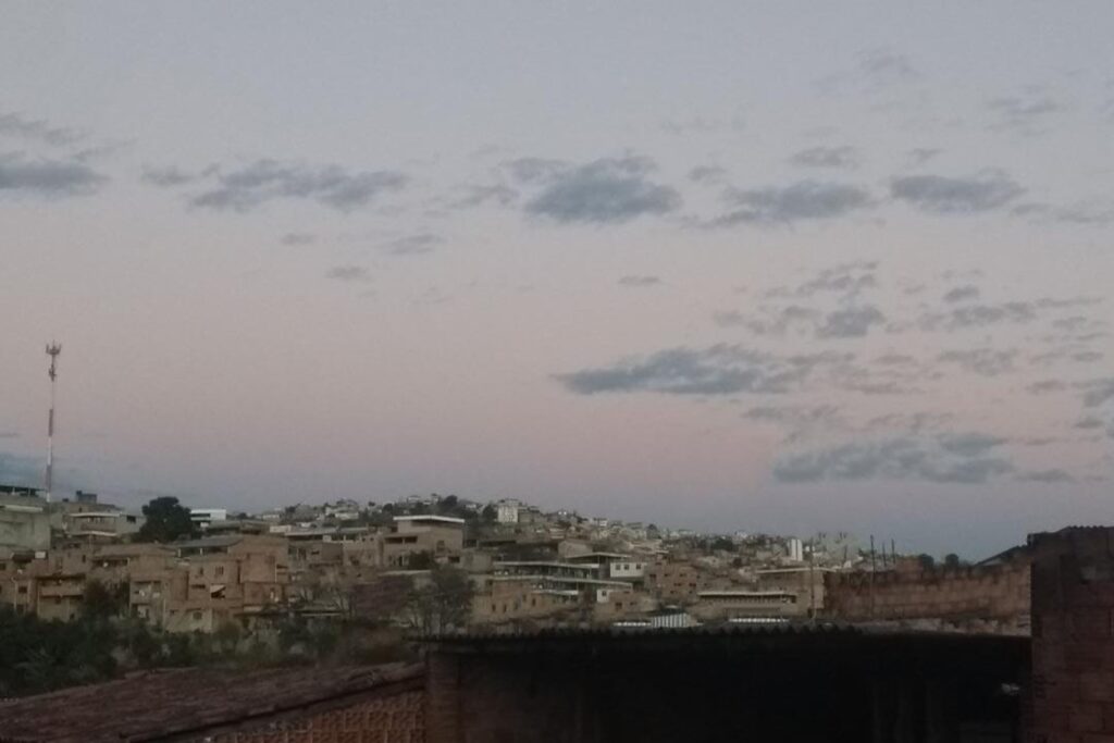 A imagem é a frente de um cartão postal, apresentando uma fotografia em cores, predominando um tom violeta. Está anoitecendo. Trata-se de uma vista panorâmica sobre um aglomerado. Em primeiro plano, na extremidade inferior da imagem, aparece o topo de algumas construções com tijolos aparentes. Em segundo plano, ainda na faixa inferior, muitas casas aglomeradas, a maioria também com tijolos aparentes. Na lateral esquerda existe uma grande e fina torre laranja e branca. Ao fundo, a maior parte do quadro é ocupado pelo céu em tons de cinza e lilás com nuances avermelhados e poucas nuvens, remetendo ao momento logo após ao pôr-do-sol. #PraCegoVer #ParaTodosVerem