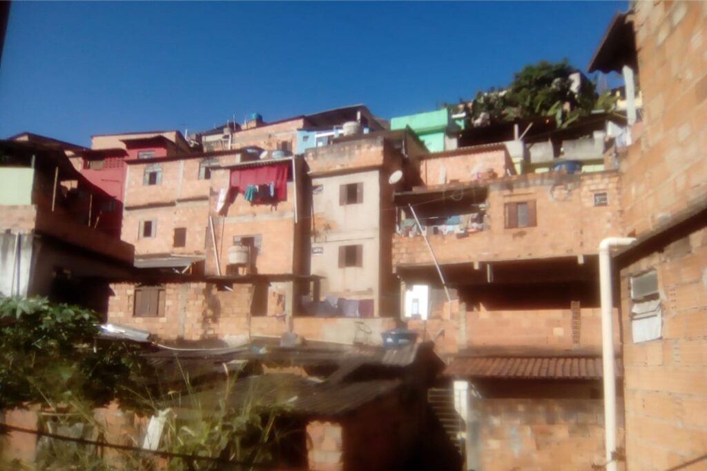 A imagem é a frente de um cartão postal, apresentando uma fotografia onde predominam tons alaranjados e um azul forte. É dia. Trata-se de uma pequena faixa de um aglomerado de casas populares verticalizadas. Em primeiro plano, no canto inferior esquerdo, uma construção mais baixa se mistura a um pouco de vegetação. Em segundo plano, em destaque, ocupando quase toda a imagem, construções de dois e três andares, de tijolos aparentes, muito próximas umas das outras. O sol forte ilumina as fachadas. Ao fundo, em toda a extremidade superior da imagem, um céu muito azul sem nuvens. #PraCegoVer #ParaTodosVerem