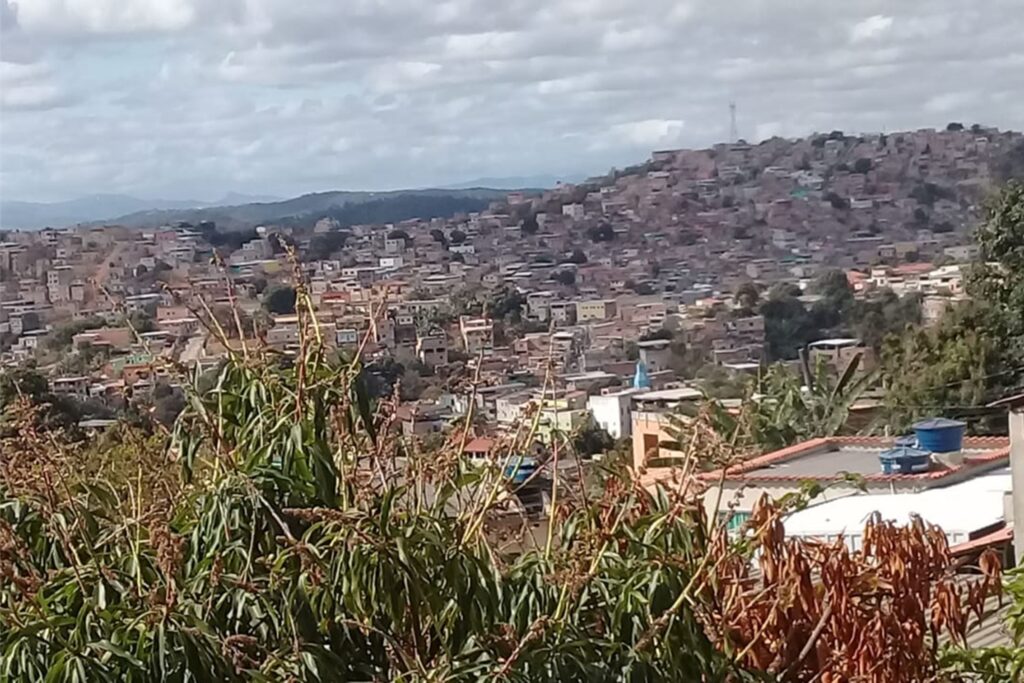 A imagem é a frente de um cartão postal, apresentando uma fotografia panorâmica onde predominam tons de cinza e um pouco de verde. O dia está nublado. Trata-se de uma paisagem panorâmica de periferia urbana, com relevo de morros. Em primeiro plano, na parte inferior, vemos a copa de uma árvore e, à direita, uma laje com duas caixas d’água azuis.  Em segundo plano, por toda extensão da paisagem, grande número de casas formam uma extensa mancha urbana. Ao fundo, há serras que já fora da mancha urbana, além do céu com muitas nuvens, que ocupa toda a parte superior da imagem. #PraCegoVer #ParaTodosVerem