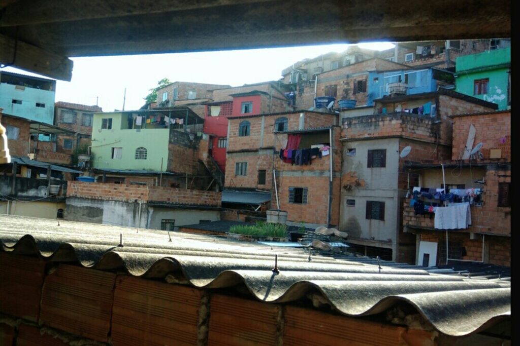 A imagem é a frente de um cartão postal, apresentando uma fotografia onde predominam tons alaranjados de tijolos.  Está de dia e o céu está nublado.  Trata-se de um aglomerado de casas populares. Em primeiro plano, na faixa superior da imagem, é possível ver apenas a extremidade interna um telhado. Em segundo plano, na faixa inferior, se vê um outro telhado completo, feito de telhas grandes e onduladas em tom cinza, iluminado pela luz do sol. Vê-se ainda a primeira camada de tijolos logo abaixo dele. Em terceiro plano, há muitas casas, de diferentes formas e tamanhos, a maioria em tijolo aparente. Algumas se destacam por estarem pintadas de cores fortes. Há muitos detalhes como diferentes tipos de janelas, roupas no varal, caixas d’água e antenas de tv. #PraCegoVer #ParaTodosVerem