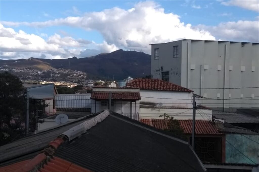 A imagem é a frente de um cartão postal, apresentando uma fotografia em cores, com predomínio do branco, cinza e marrom. O dia está claro. Trata-se de uma serra vista acima dos telhados. Em primeiro plano, um telhado cinza escuro. Em segundo plano, do outro lado da rua, outros telhados, de diferentes materiais, vistos de cima. Em terceiro plano, no canto direito, um grande galpão pintado de cinza claro, bem alto em relação às outras construções ao redor. Ao fundo, com exceção da faixa direita que está encoberta pelo galpão, se vê ao longe a continuação da mancha urbana e uma serra imponente se destaca no horizonte. Na faixa superior, céu azul com nuvens brancas. #PraCegoVer #ParaTodosVerem