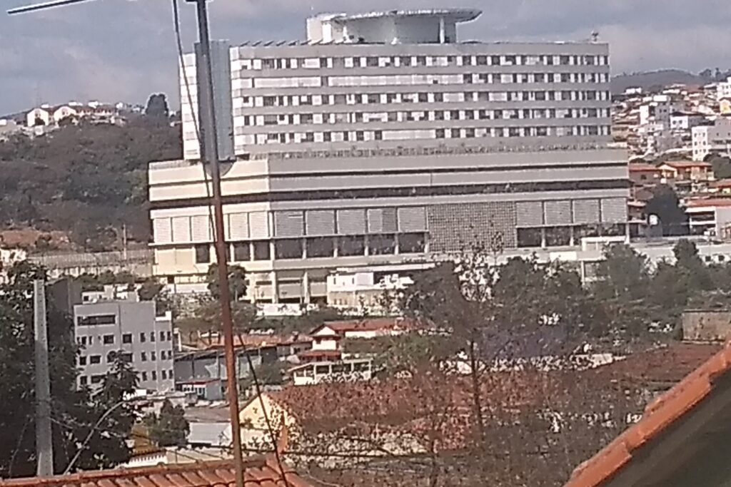 A imagem é a frente de um cartão postal, apresentando uma fotografia onde predominam tons de branco e terrosos. O dia está nublado. Trata-se de um grande prédio em destaque na paisagem urbana. Em primeiro plano, ao centro, vê-se uma antena e o topo de algumas árvores e telhados. Em segundo plano, em destaque, o prédio cinza, largo e retangular, com muitas janelas. Ao fundo, outros prédios menores, casas, montanhas e o céu nublado. #PraCegoVer #ParaTodosVerem