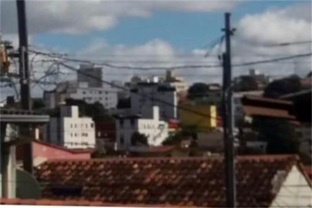 A imagem é a frente de um cartão postal, apresentando uma fotografia em cores, com predomínio do branco e marrom. A imagem está fora de foco. O dia está claro. Trata-se de uma paisagem urbana.  Em primeiro plano, dois postes de eletricidade ligados por fios. Em segundo plano, ocupando quase toda a faixa inferior da imagem, o topo de um telhado colonial. Em terceiro plano, algumas camadas de casas e pequenos edifícios ocupam um terreno ligeiramente elevado. Ao fundo, o céu está azul com muitas nuvens. #PraCegoVer #ParaTodosVerem