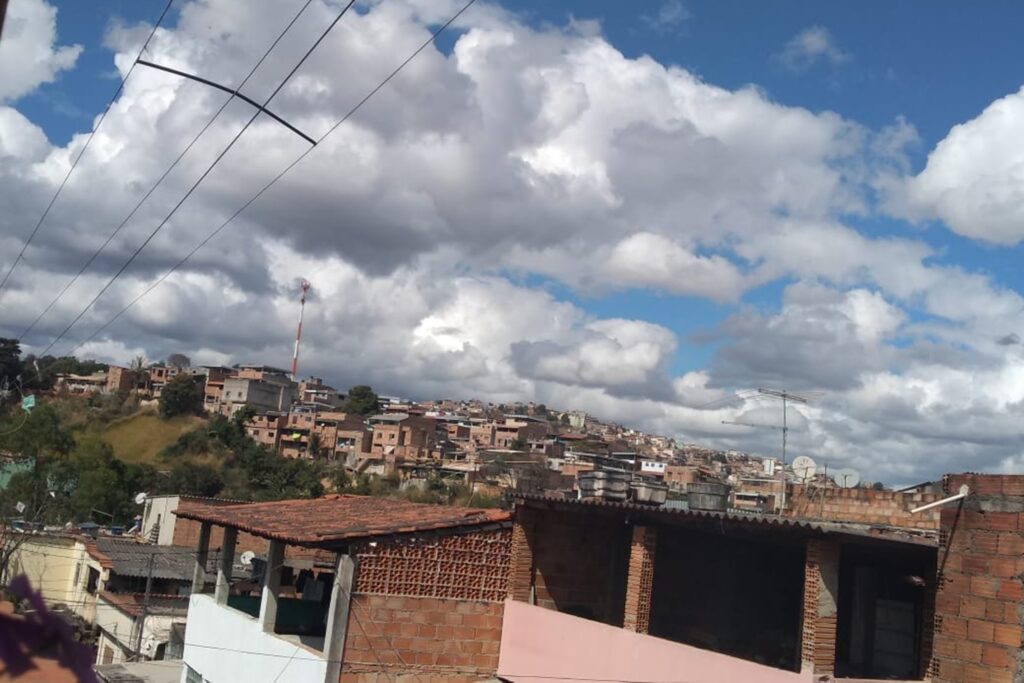 A imagem é a frente de um cartão postal, apresentando uma fotografia em cores onde predominam tons terrosos. O dia está claro. Trata-se de uma visão panorâmica de um aglomerado de casas. Em primeiro plano, na faixa superior, quatro fios de energia se destacam no canto superior esquerdo. Na faixa inferior, destacam-se as coberturas de duas casas em uma ladeira. Uma delas, com roupas no varal. Em segundo plano, na faixa inferior, há muitas casas, muito próximas umas das outras, quase todas em tijolo aparente. No canto esquerdo, uma pequena área verde com vegetação rasteira e algumas árvores. Ao fundo, ocupando toda a metade superior da imagem, o céu parcialmente nublado, tem manchas azuis e nuvens densas. #PraCegoVer #ParaTodosVerem