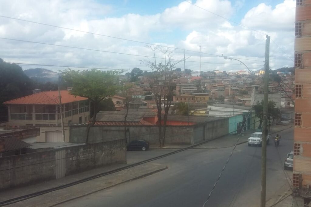 A imagem é a frente de um cartão postal, apresentando uma fotografia em cores onde predominam tons de cinza. O dia está nublado. Trata-se de uma paisagem urbana. Em primeiro plano, na extremidade direita, a imagem é emoldurada por uma parede de tijolos aparentes em tons alaranjados e próxima a ela está um poste de iluminação da rua. Em segundo plano, se vê uma esquina de ruas asfaltadas, onde circulam carros e uma moto. Em terceiro plano, estão dois quarteirões de casas cercadas por muros com calçadas com poucas árvores. Uma das casas, no canto esquerdo, se destaca por ser grande, circundada por varanda de vidro e com telhado colonial. Ao fundo, se vê muitas casas e o céu está coberto de nuvens brancas, com pequenas faixas azuis. #PraCegoVer #ParaTodosVerem