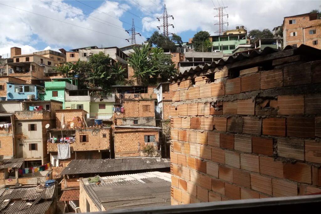 A imagem é a frente de um cartão postal, apresentando uma fotografia onde predominam tons terrosos de tijolos. O dia está claro. Trata-se de um aglomerado de casas populares. Em primeiro plano, à direita, parede de tijolos aparentes, metade à sombra, metade iluminada pelo sol. Em segundo plano, outras casas, a maioria também de tijolos aparentes e algumas pintadas em tons de verde e azul. Entre as casas, detalhes como roupas no varal e caixas d'água.  Ao fundo, na parte superior da imagem, céu azul com nuvens brancas, três antenas de transmissão de energia e algumas árvores misturadas às casas. #PraCegoVer #ParaTodosVerem