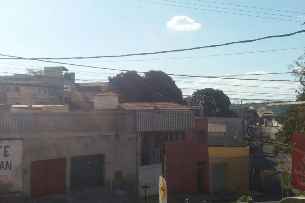 A imagem é a frente de um cartão postal, apresentando uma fotografia em cores. O dia está ensolarado. Trata-se de uma rua com casas coloridas. Em primeiro plano, muitos fios elétricos atravessam a imagem na horizontal. Em segundo plano, se destacam três fachadas de casas, todas quadradas com portões de garagem. A que está mais à esquerda é revestida de cimento, com portões vermelho e azul. A seguinte está pintada de vermelho com portão grande e branco e uma porta pequena. A terceira fachada é amarela, com portão cinza claro. Ao fundo se vê outras casas e a copa de algumas árvores. No canto inferior direito, a continuação da rua após uma descida com curva suave para a esquerda. Na faixa superior, o céu está claro, com poucas nuvens. #PraCegoVer #ParaTodosVerem