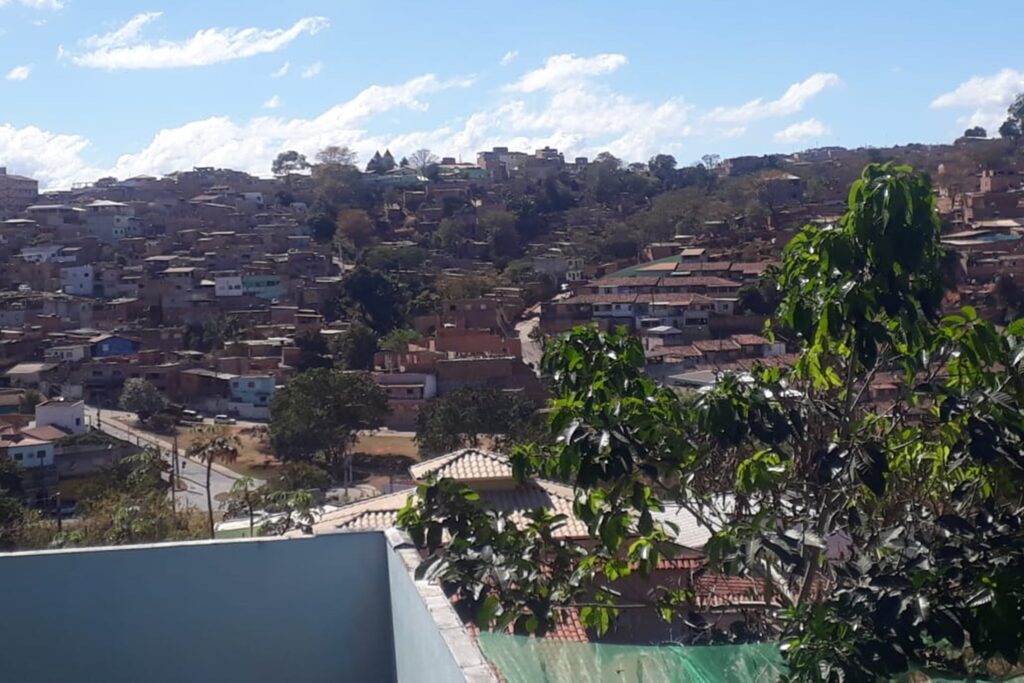 A imagem é a frente de um cartão postal, apresentando uma fotografia colorida em que predominam tons de azul, verde e marrom. O dia está claro. Trata-se de quarteirões de um bairro residencial. Em primeiro plano, na faixa inferior, à direita, uma árvore pequena e, à esquerda, a quina de um muro azul baixo. Em segundo plano, na faixa central, se vê muitas casas se estendendo até o alto de uma colina. A maior parte está aglomerada do lado esquerdo e, entre elas, há algumas árvores e ruas. Ao fundo, na faixa superior, o céu é azul com poucas nuvens. #PraCegoVer #ParaTodosVerem