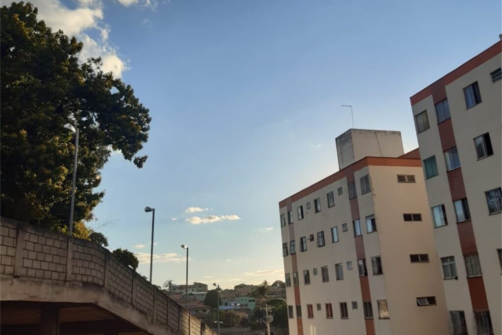 A imagem é a frente de um cartão postal, apresentando uma fotografia colorida em que predominam tons claros de bege e azul. É dia. A imagem está levemente angulada para cima, mirando o céu visto de um conjunto habitacional. Em primeiro plano, à direita, há dois prédios, com mesmo formato e acabamento, com muitas janelas e pintados de bege com faixas marrons. Na porção esquerda da imagem, um muro de tijolos cinzas protege uma rampa para carros. Em segundo plano, no canto esquerdo, por detrás do muro, há uma grande árvore folhosa. Ao fundo, o céu predomina na maior parte do quadro, azul, muito iluminado e com poucas nuvens. #PraCegoVer #ParaTodosVerem