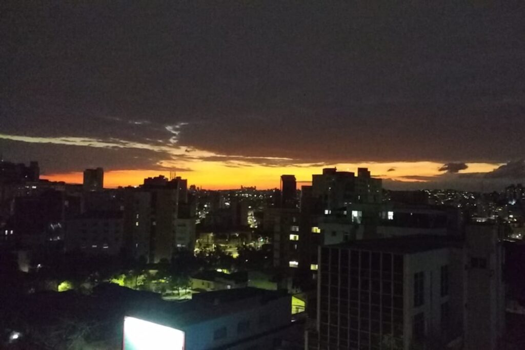 A imagem é a frente de um cartão postal, apresentando uma fotografia em que predomina o preto e se destaca o laranja. É início da noite. Trata-se de paisagem urbana. Em primeiro plano, na faixa inferior, vários prédios com algumas luzes acesas. Em segundo plano, a continuação da cidade como pontos luminosos. Ao fundo, no horizonte, uma faixa alaranjada de luz remanescente e, acima, o céu escuro. #PraCegoVer #ParaTodosVerem