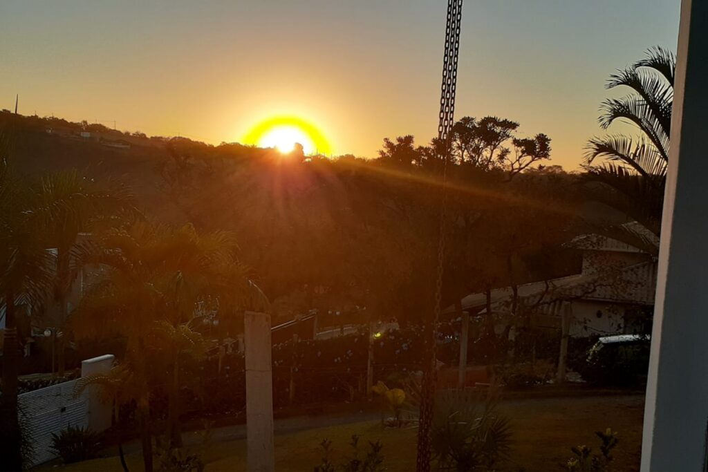 A imagem é a frente de um cartão postal, apresentando uma fotografia onde predominam tons amarelados. É fim de tarde. Trata-se do pôr-do-sol por detrás de uma colina. Em primeiro plano, correntes finas cortam a imagem na vertical e, no canto direito, há a moldura branca de uma janela. Em segundo plano, o gramado de um jardim é protegido por cercas. Em terceiro plano, a rua com algumas casas e árvores. Ao fundo, o sol se põe em evidência no centro da foto. Há um arco em forte tom de amarelo em seu entorno e seus raios se projetam sobre a paisagem. #PraCegoVer #ParaTodosVerem