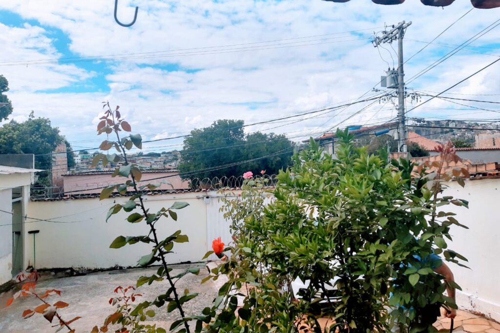 A imagem é a frente de um cartão postal, apresentando uma fotografia colorida em que predomina o branco. É dia. Trata-se de um quintal com garagem a céu aberto. Em primeiro plano, há um arbusto verde, misturado a uma roseira com rosas vermelhas e cor de rosa. Por entre as plantas se entrevê parte do corpo de uma pessoa segurando uma mangueira de água e de um automóvel branco. Em segundo plano, há um pátio com chão de cimento, cercado por muro branco com portão de garagem. Em terceiro plano, para além do muro, se vê o topo de casas e a copa de algumas árvores. Na faixa superior, céu azul com muitas nuvens brancas. #PraCegoVer #ParaTodosVerem