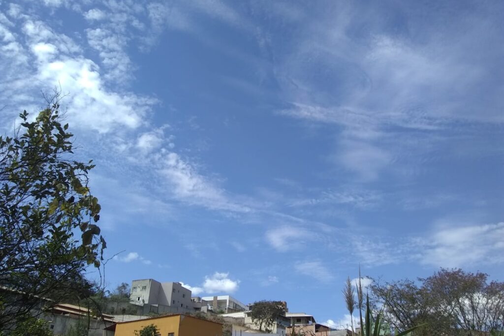 A imagem é a frente de um cartão postal, apresentando uma fotografia colorida em que predomina o azul do céu diurno. Em primeiro plano, a borda inferior e parte da lateral esquerda são emolduradas pelas copas de algumas árvores e pelo topo de algumas casas e prédios. Em segundo plano, quase toda a imagem apresenta o céu azul, com nuvens muito suaves, de consistência rarefeita. #PraCegoVer #ParaTodosVerem