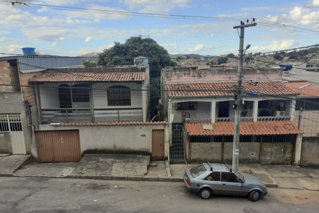 A imagem é a frente de um cartão postal, apresentando uma fotografia colorida em que predominam tons cinzas. O dia está claro. A foto está levemente angulada de cima para baixo, registrando a fachada de duas casas. Em primeiro plano, há uma rua com um carro cinza, de modelo antigo, estacionado. Em segundo plano, a calçada com um poste de eletricidade e os muros das duas casas com portões de garagem e para pedestres. Em terceiro plano, por trás dos muros e elevadas em relação a eles, é possível ver as varandas das casas. Em cada uma, uma porta, duas janelas e telhados coloniais. Em quarto plano, pelo estreito espaço entre as casas e acima delas, vê-se uma grande árvore, muito folhosa. Bem ao fundo, à direita da imagem, outras casas e, à esquerda, uma serra. Na faixa superior, céu azul com nuvens brancas. #PraCegoVer #ParaTodosVerem